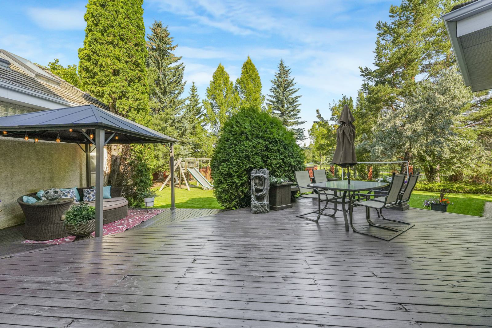 The upper deck under its covered lounge, dining table set out, looking across the lawn to mature trees.