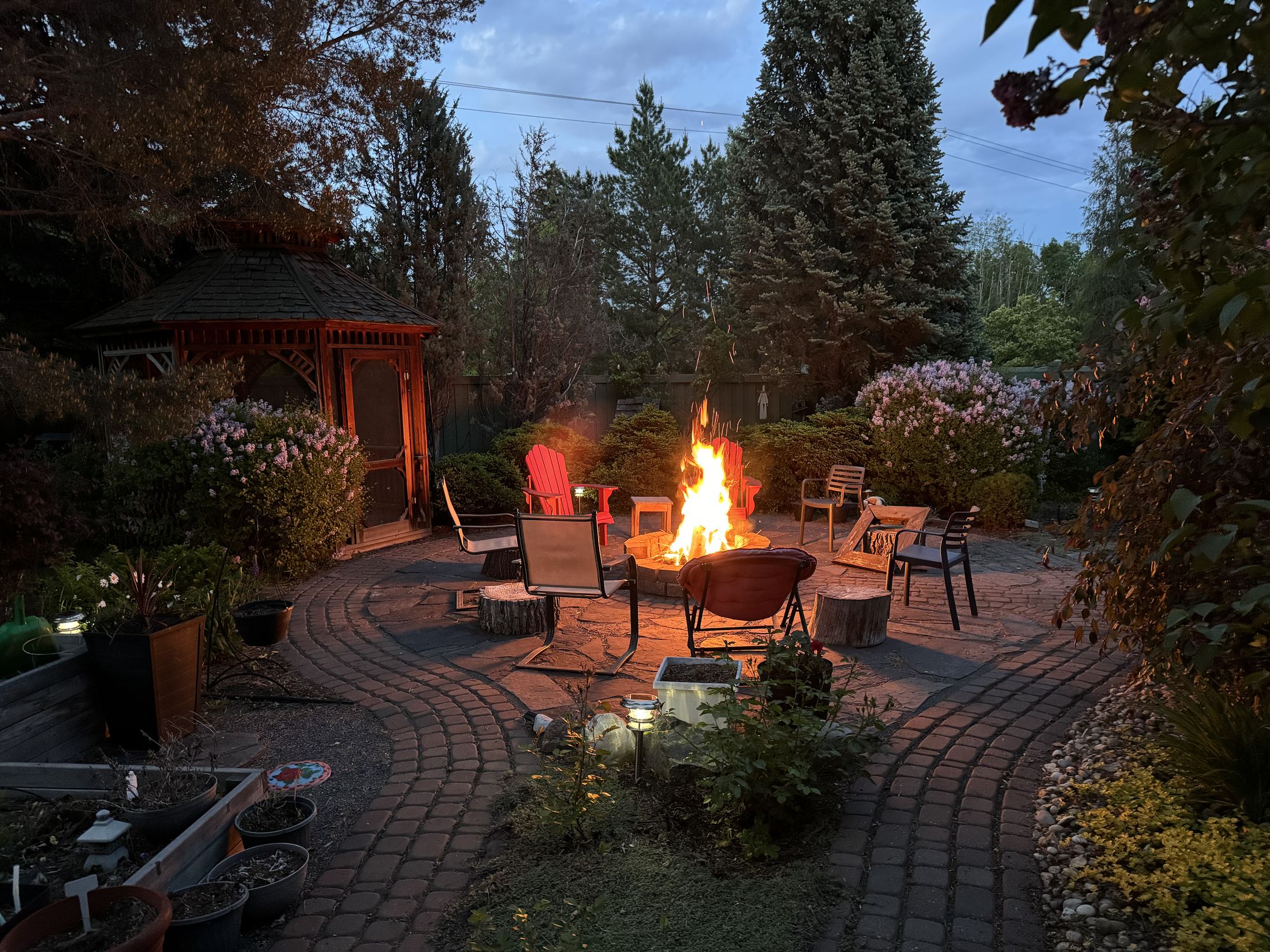 The flagstone fire pit terrace at dusk, fire lit, ringed by chairs with the gazebo and mature trees behind.