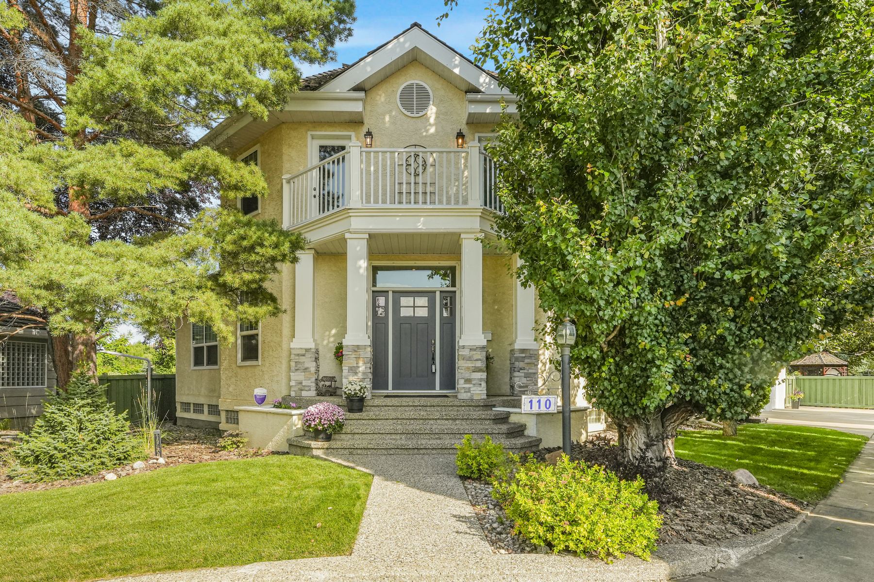 The front elevation of 110 Weaver Drive in summer, stucco and stone with a columned portico and a balcony above.