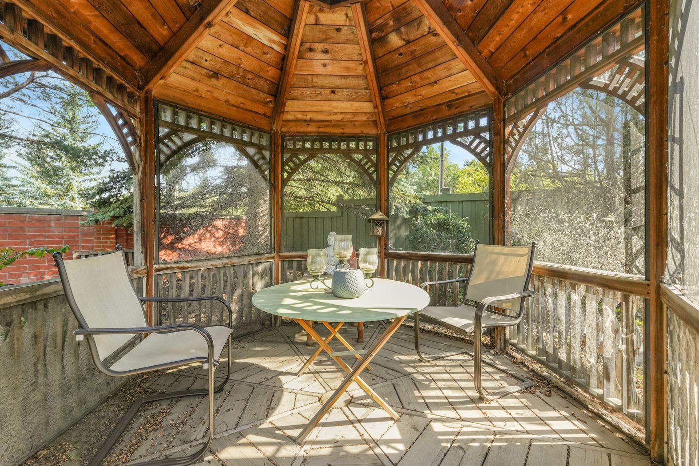 Inside the screened cedar gazebo, a table set for two under the timbered roof with the garden beyond.