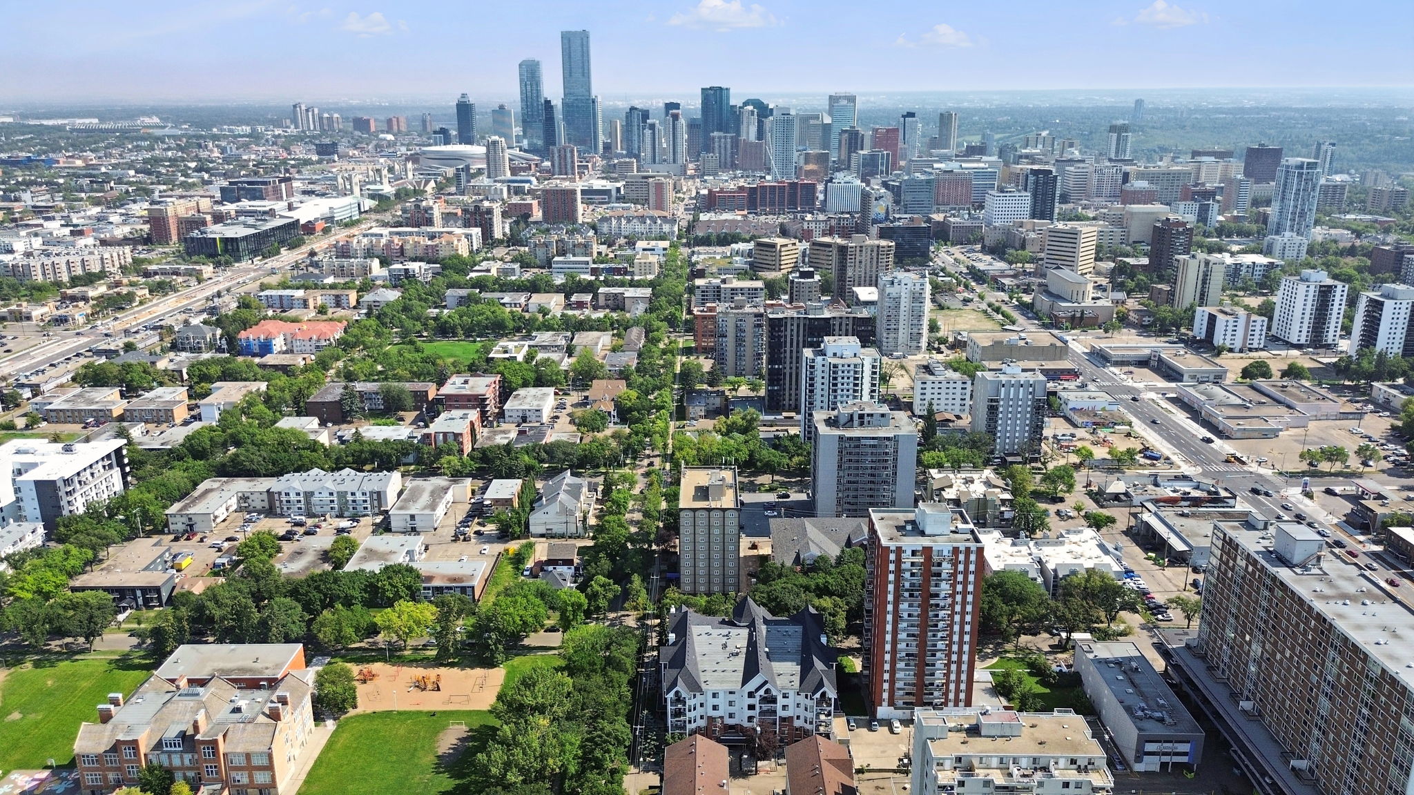 Aerial view over Wîhkwêntôwin toward the downtown Edmonton skyline in summer, tree-lined streets and mid-rise buildings in the foreground.