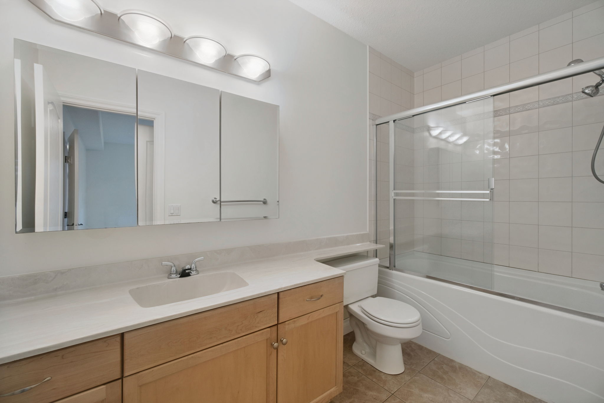 The four-piece ensuite with a maple vanity, a wide mirror under a strip of lights, and a tub with a glass shower door.