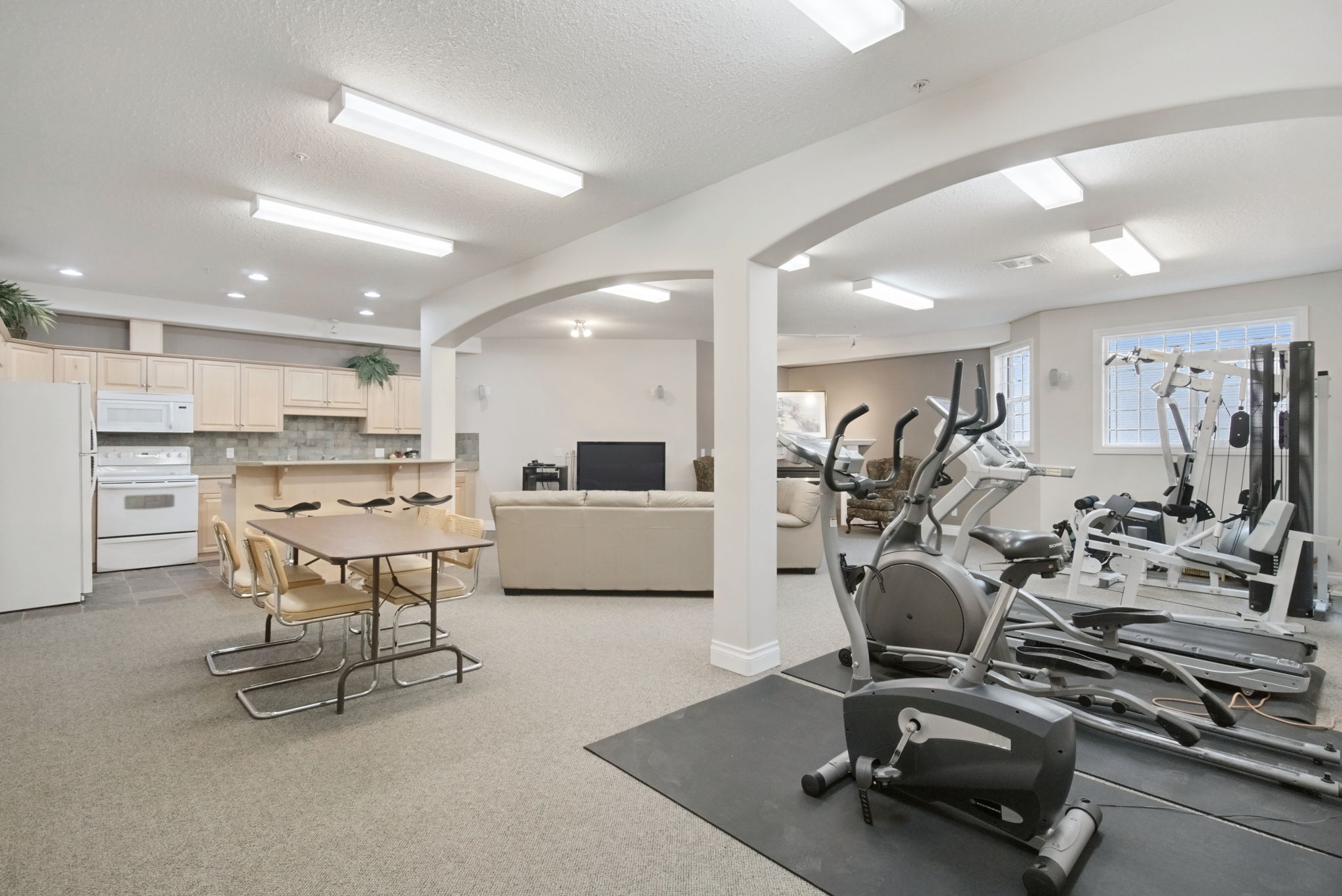 The exercise room with a treadmill, elliptical, bikes and a weight machine on a rubber floor beside the social room.