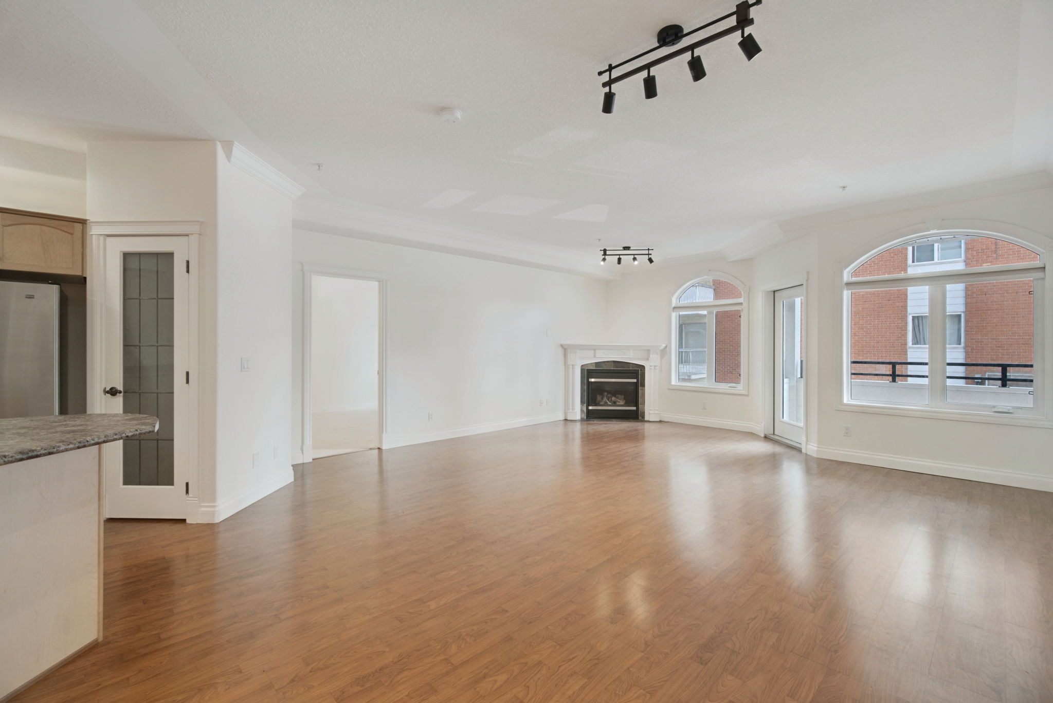 The living room of unit 203, an open space under nine-foot ceilings with a gas fireplace, two arched windows and a door to the balcony, wood-look laminate floors running through to the kitchen.
